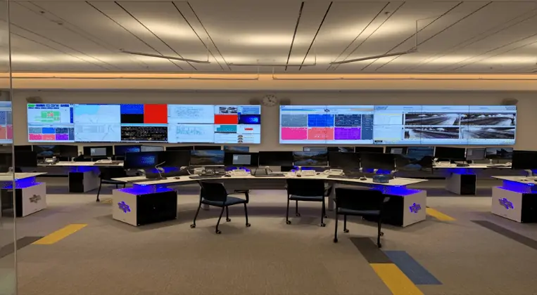 Monitorreihen im Büro einer Leitstelle mit verschiedensten Grafiken und Daten über Versorgung und Transportation abgebildet. Im Vordergrund steht ein Schreibtisch mit drei Stühlen.